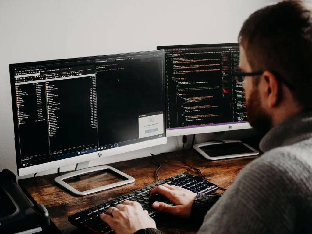 Developer typing code on dual monitors at a wooden desk Website development services: developer typing code on dual monitors at a wooden desk